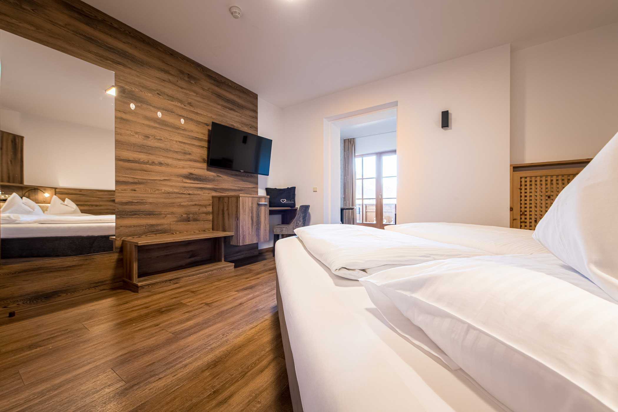 Modernes Hotelzimmer mit Holzakzenten, einem Doppelbett, einem an der Wand montierten Fernseher, einem Schreibtisch und einem großen Fenster mit Balkonblick. - Hotel Waldheim