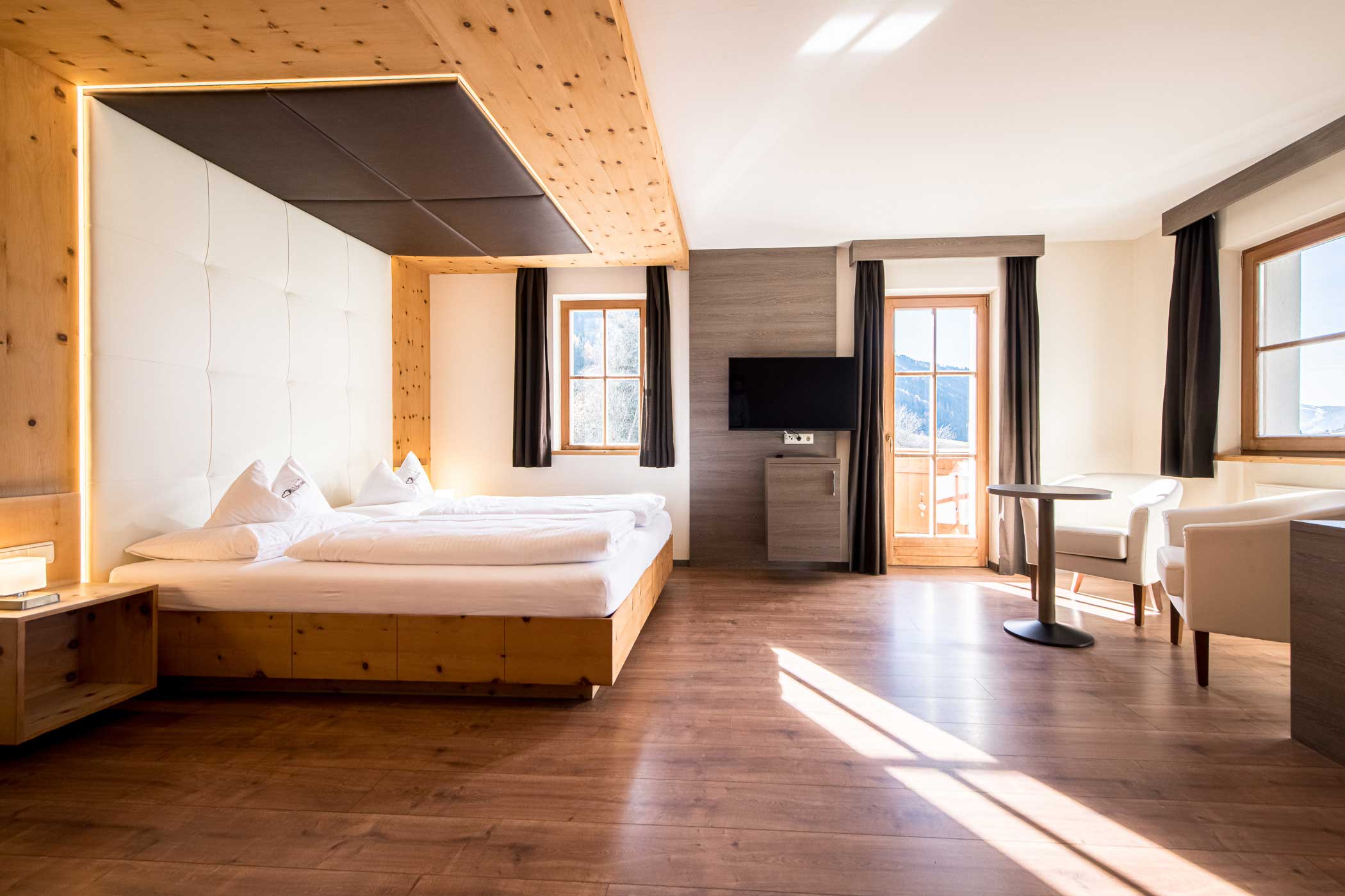 Helles Hotelzimmer mit Holzakzenten, einem großen Bett, TV, Sitzecke und Balkon mit Bergblick. - Hotel Waldheim