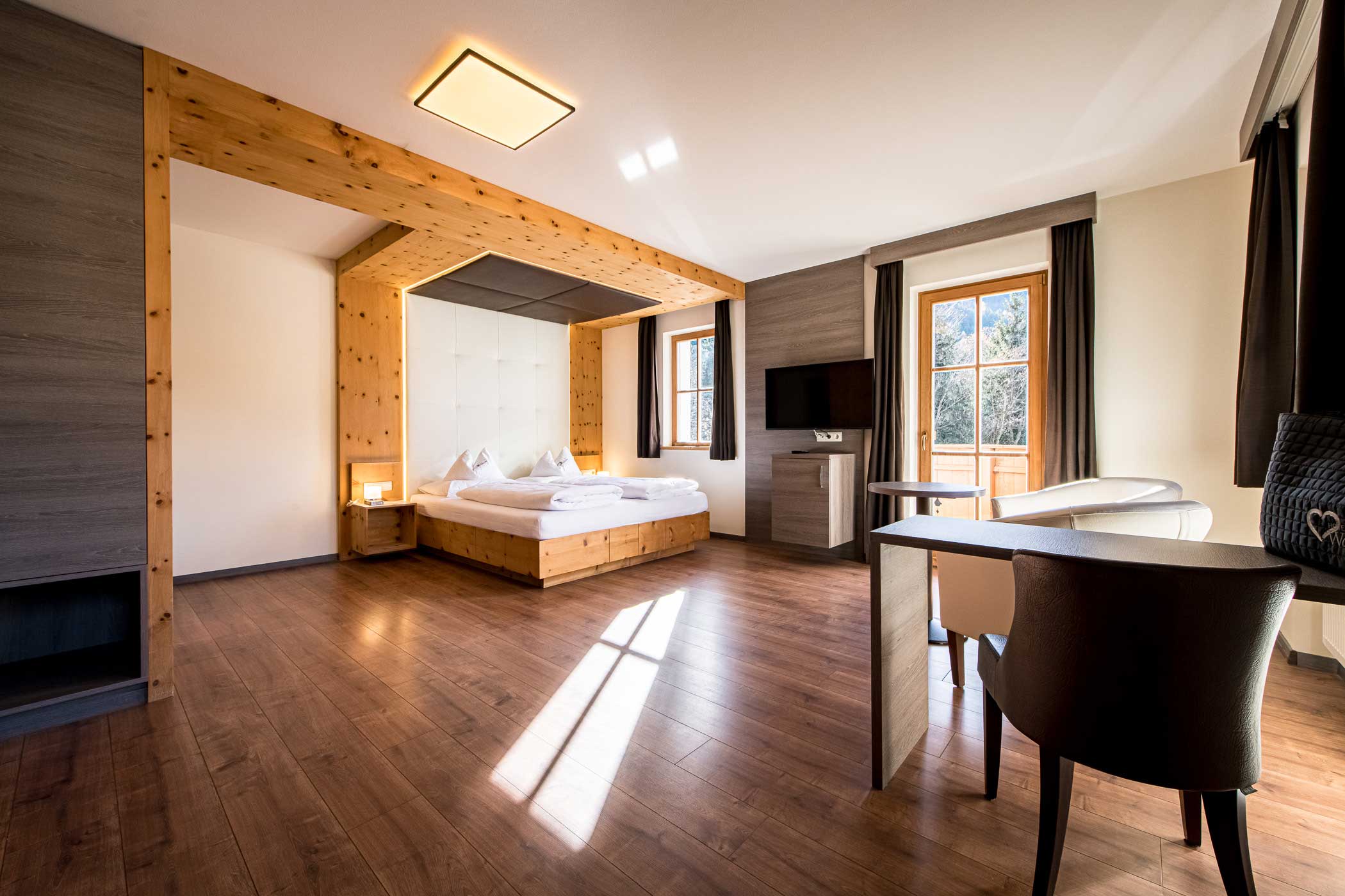 Modernes Hotelzimmer mit Holzboden, großem Bett, Sonnenlicht, TV, Schreibtisch und zwei Stühlen am Fenster. - Hotel Waldheim
