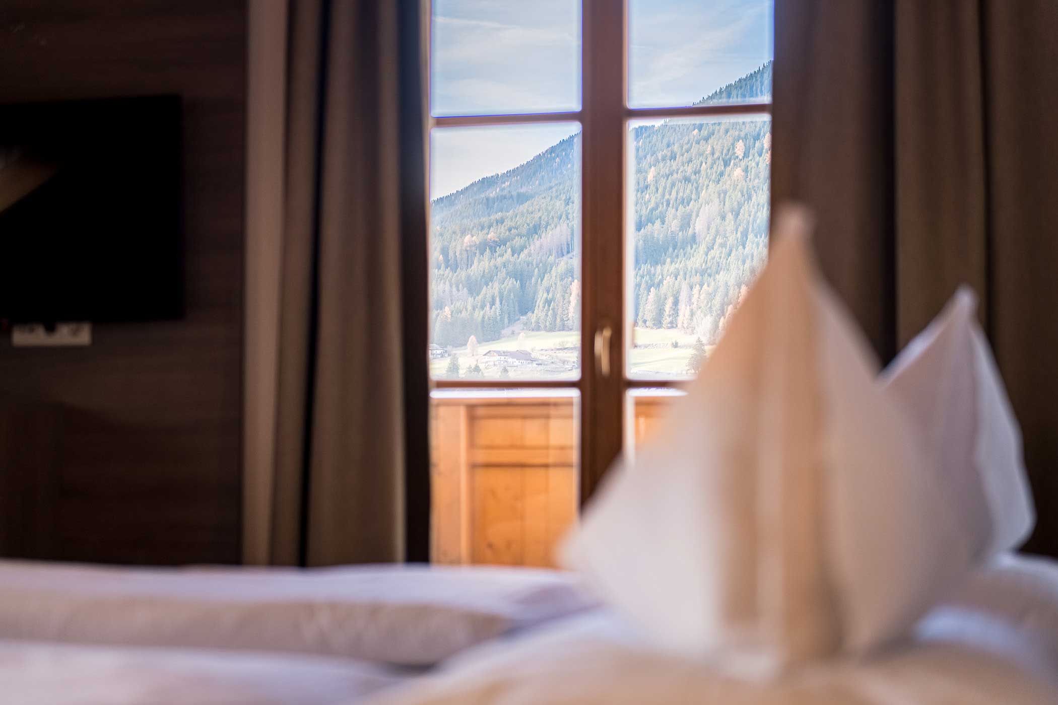 Ein gemütliches Bett mit Blick auf die Berge durch ein Fenster mit Vorhängen in einem sanft beleuchteten Raum. - Hotel Waldheim