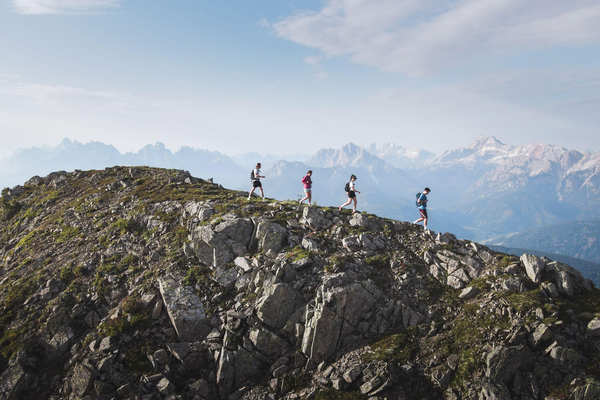 Vier Personen laufen entlang eines felsigen Bergrückens mit fernen Gipfeln und blauem Himmel im Hintergrund. - Hotel Waldheim