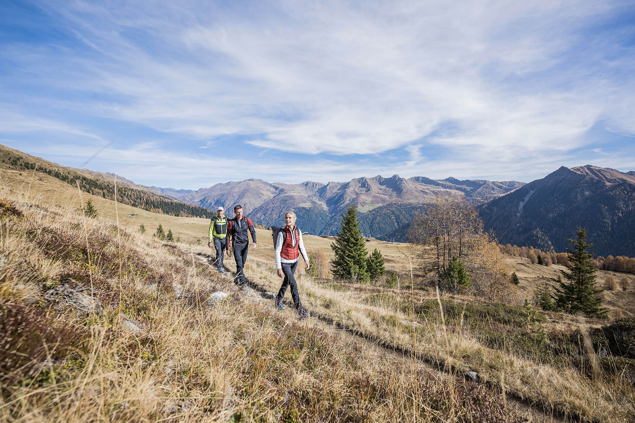 Drei Personen wandern auf einem grasbewachsenen Bergpfad mit malerischen Bergen und blauem Himmel im Hintergrund. - Hotel Waldheim
